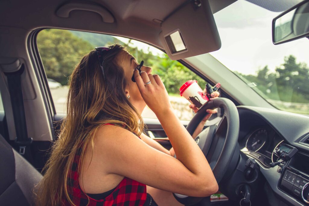 Woman applying mascara while sitting in the driver&rsquo;s seat of a car, holding a coffee cup, illustrating distracted driving behavior