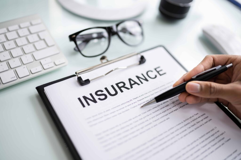 Hand holding pen over insurance policy document on clipboard, with glasses and keyboard on desk, representing insurance coverage and policy review