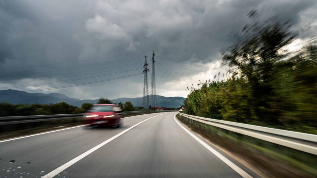 Red car driving fast on a curving highway under dark storm clouds, motion blur showing speed, rural landscape and power lines in the background